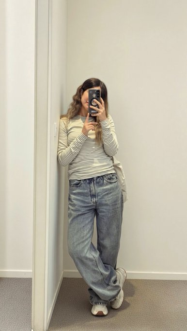 A woman takes a mirror selfie wearing a striped long-sleeve shirt, loose blue jeans, and chunky white sneakers.