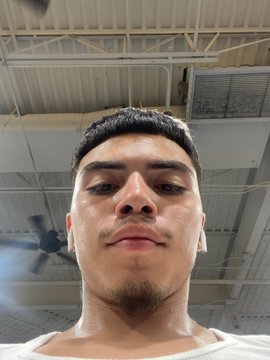 A young man with short dark hair and a light goatee takes a close-up selfie from a low angle, his face glistening with sweat against the backdrop of an industrial ceiling with exposed pipes, a ceiling fan, and metal beams. The "#htx" hashtag suggests the photo was taken in Houston, Texas, possibly after a workout or sports activity given his perspiration and the gym-like setting visible overhead.