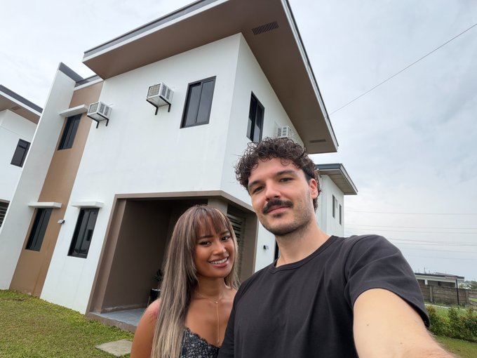 A selfie of a smiling couple standing in front of a modern white two-story house with multiple air conditioning units mounted on the exterior walls.