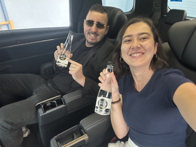Selfie-style photo of a smiling man in sunglasses and a woman in a navy blue top sitting in the back seat of a vehicle, both holding up small bottles of water in a celebratory toast.