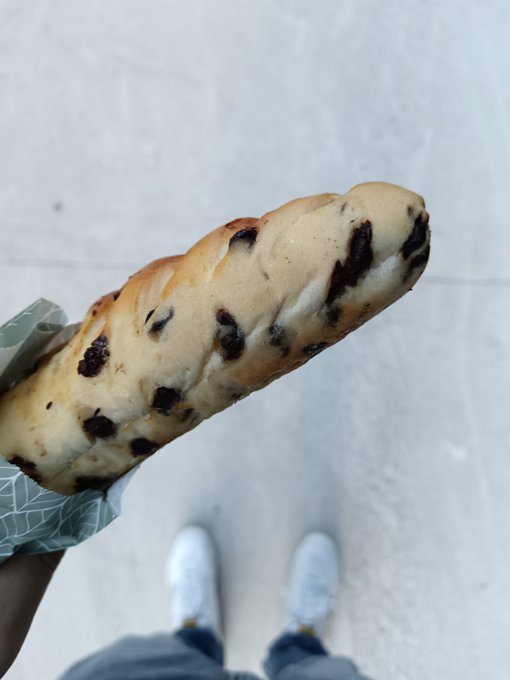 A first-person view looking down at a hand holding a large chocolate chip pastry, with the photographer's white sneakers and pants visible against a light gray floor.