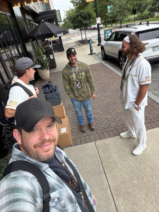 A selfie taken by a man in a black cap showing three other men, including one in a white Oakland Athletics jersey, standing on a sidewalk outside a restaurant.