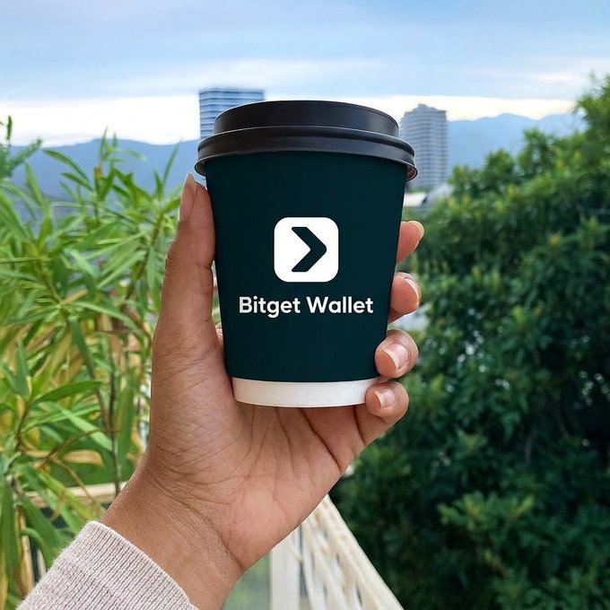 A hand holding a dark green coffee cup featuring the 'Bitget Wallet' logo with a white arrow icon, set against a backdrop of greenery and distant buildings.
