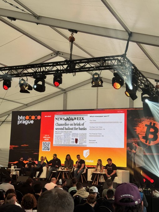 The image shows a group of people sitting around a table, with a large screen displaying information about Bitcoin. There are at least 12 people visible in the scene, some seated while others stand or sit on chairs arranged around the table. The audience appears engaged as they listen and observe the presentation. The post related to this event reads: "Satoshi Millionaire packed as always".