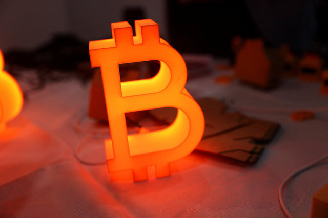 A physical, 3D "B" Bitcoin logo illuminated in orange and yellow light sits on a desk next to paper cutouts and cables.