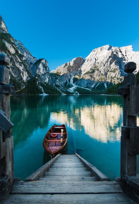 A serene landscape photograph of a wooden boat moored at a wooden staircase leading down to a crystal-clear turquoise lake, surrounded by dramatic rocky mountains under a clear blue sky.