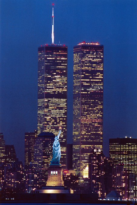 A nighttime cityscape photograph showing the illuminated Twin Towers of the World Trade Center behind the Statue of Liberty.
