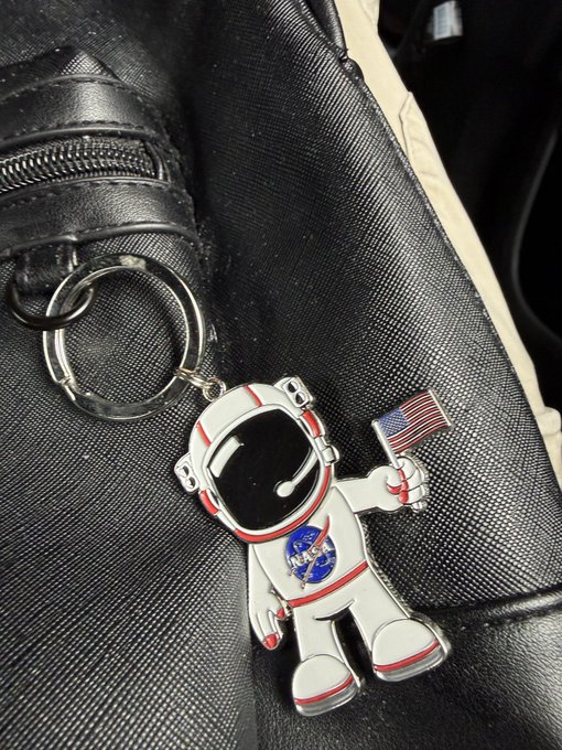A metal keychain featuring an astronaut figure with a NASA logo and a small American flag, attached to a keyring and resting on a black leather bag.