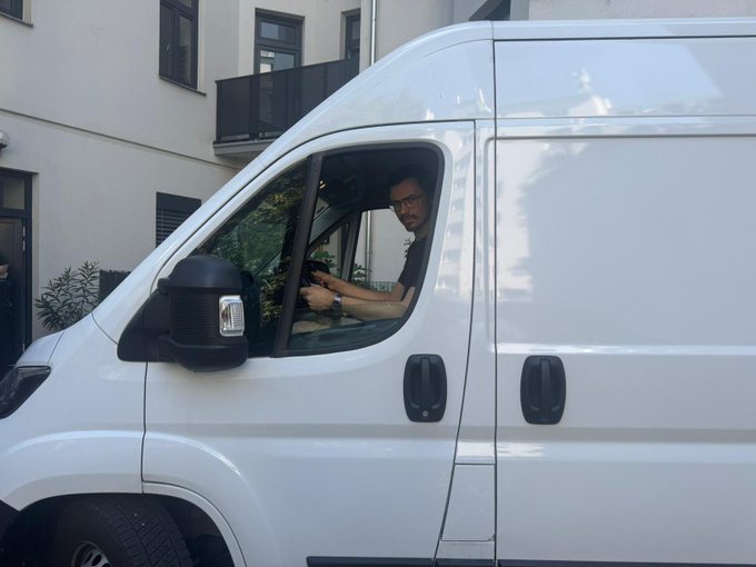 A man wearing glasses and a dark shirt sits in the driver's seat of a white van, looking out the open window at the camera.