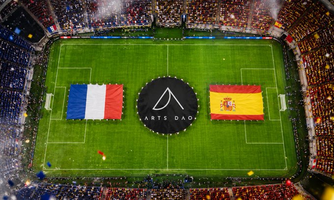 An aerial view of a packed soccer stadium features large French and Spanish flags on the field, with a black circular 'ARTS DAO' banner in the center circle.