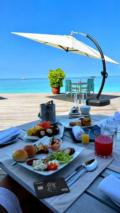 The image shows a table set up with plates of food, glasses filled with juice, and an umbrella providing shade from the sun. The breakfast spread includes items such as bread, fruit, and other dishes. A person can be seen sitting at the table enjoying their meal under the umbrella. The post features a description of a beautiful ocean view, suggesting that this location is likely near the beach or by the water's edge. The image also contains text about spending seamlessly with cryptocurrency, which could indicate that the breakfast spot offers a unique dining experience where guests can pay for their meals using cryptocurrency instead of traditional payment methods.