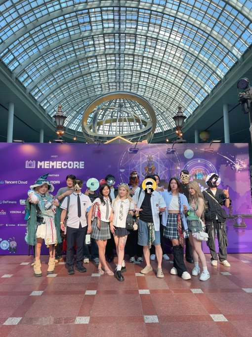 A group of cosplayers and attendees pose together at a 'MEMECORE' event, with various sponsor logos including Tencent Cloud and DeFiware visible on the purple backdrop.