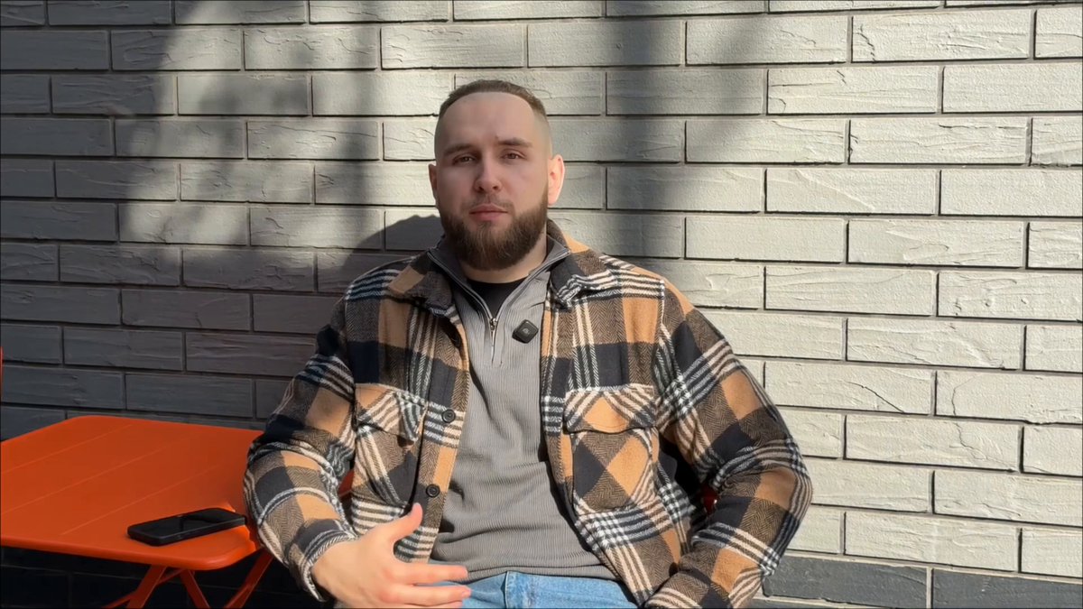 The image features a man sitting at an orange table with his legs crossed while wearing a plaid shirt. The background of the photo shows a brick wall, which adds to the overall aesthetic of the scene. There are also two cell phones visible in the image - one on the table and another placed nearby.