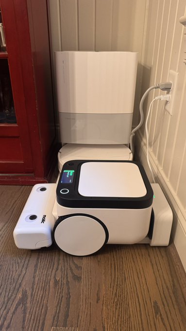 A compact white Matic robot vacuum sits docked at its slim charging station against a paneled wall on a hardwood floor, demonstrating the brand's notably narrow and space-saving design. The setup illustrates how the robotic vacuum's small footprint allows it to fit neatly into tight corners of a home.