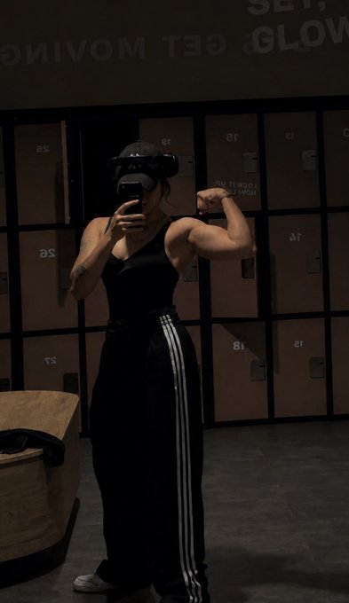 A woman takes a mirror selfie wearing a black tank top, black track pants with white stripes, and a VR headset, while flexing her bicep in front of lockers labeled with letters and numbers.