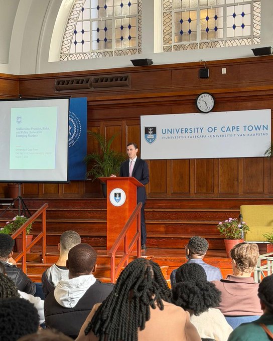 A man in a suit speaking at a podium labeled 'UNIVERSITY OF CAPE TOWN' during an event with a presentation slide titled 'Walkminster, Precinct, Risks and Bullies: Outlook for Emerging Markets'.