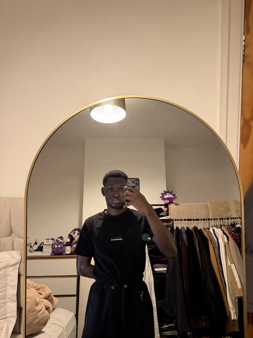 A selfie taken in a mirror showing a young man wearing a black t-shirt and shorts, holding a smartphone, with a closet full of clothes visible in the reflection behind him.