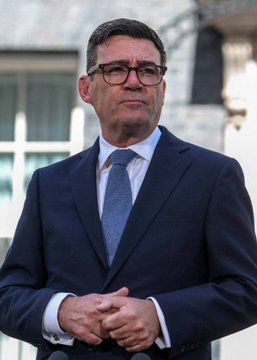 Photograph of a man in a dark suit, blue shirt, and patterned blue tie, wearing glasses, with his hands clasped in front of a blurred building background.