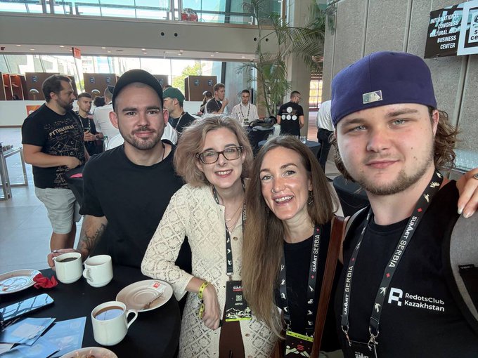 A group selfie of four people at what appears to be a convention; the person on the right wears an MLB baseball cap and a lanyard for "RedotsClub Kazakhstan."