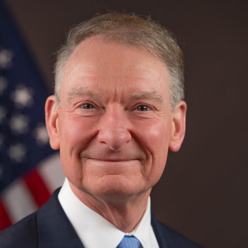 An official headshot portrait of a smiling older man in a dark suit and blue tie, with an American flag visible in the background.