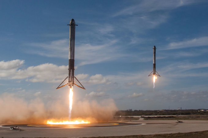 Two SpaceX Falcon rockets land simultaneously on a landing pad, with flames and smoke billowing from the one in the foreground.