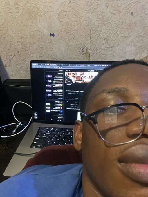 A close-up selfie of a person wearing glasses, with a MacBook in the background displaying a Discord chat window.