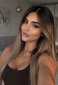 A close-up selfie of a young woman with long brown highlighted hair wearing a black top and a small cross necklace, in what appears to be an indoor setting.