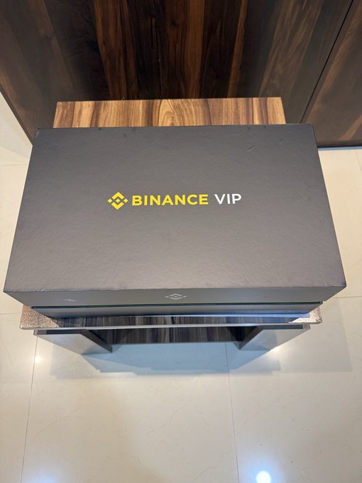 A dark gray premium box labeled 'BINANCE VIP' in gold lettering, resting on a wooden table against a wood-paneled wall.