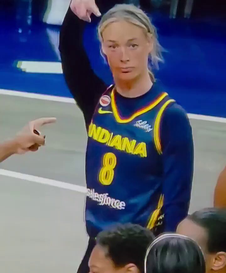 The image shows a female basketball player wearing an Indiana jersey, standing on a court with her arm raised high above her head. She appears to be celebrating or acknowledging someone by pointing at them. The background of the photo reveals that there are other people present in the scene as well.