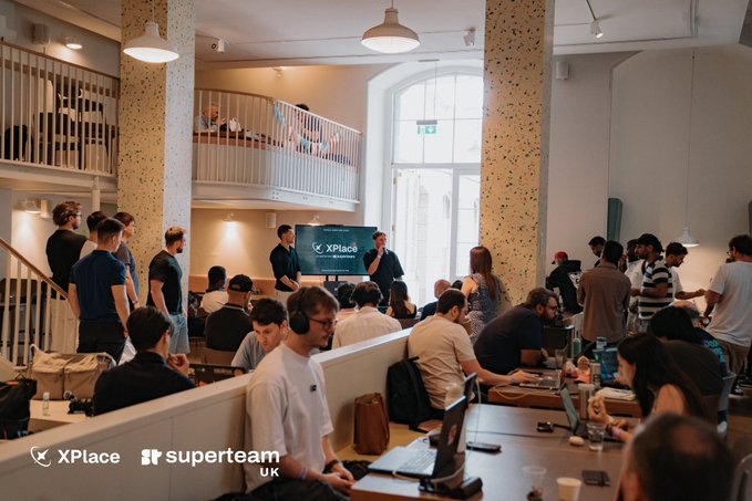 A photograph of a packed co-working or event space with people seated at long tables working on laptops, with a presentation screen visible; logos for 'XPlace' and 'superteam UK' appear in the bottom corners.