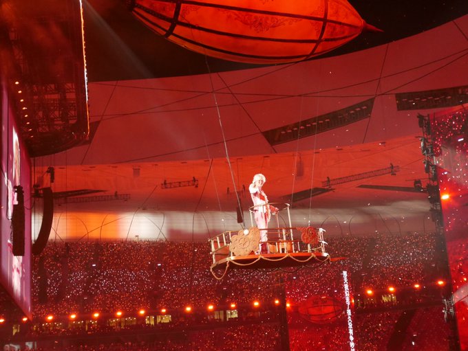 A dramatic concert scene bathed in red lighting, showing a performer on a suspended platform above a massive crowd, with large spherical structures hanging from the venue's ceiling.