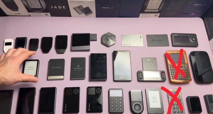 A collection of over 40 different cryptocurrency hardware wallets, including brands like Jade, SafePal X1, Ellipal, and Tangem, is displayed on a pink surface, with a person's hand visible on the left. Three devices are marked with red X's, including one in the second row resembling a Coldcard, illustrating the post's point about unexpected failure and urging forgiveness for those who previously recommended the brand.