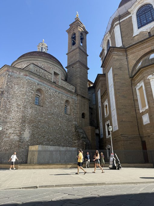 A street-level view shows an old stone building with a dome and bell tower on the left, and a more ornate facade on the right, with people walking by on the sunny pavement.