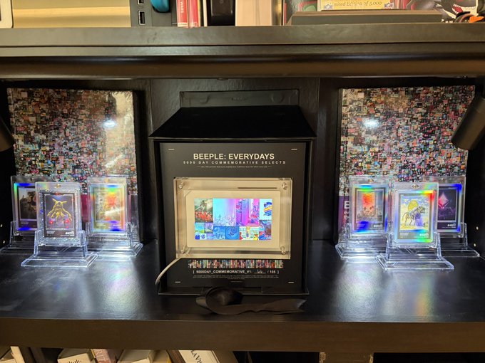 The image features a television set with a DVD player, surrounded by several framed photos of Beeple's 5001 day selections. The TV is placed on top of a shelf or display case, which also holds other items such as trading cards. The post about the trading card collection suggests that this setup might be related to the world of video games and gaming memorabilia.