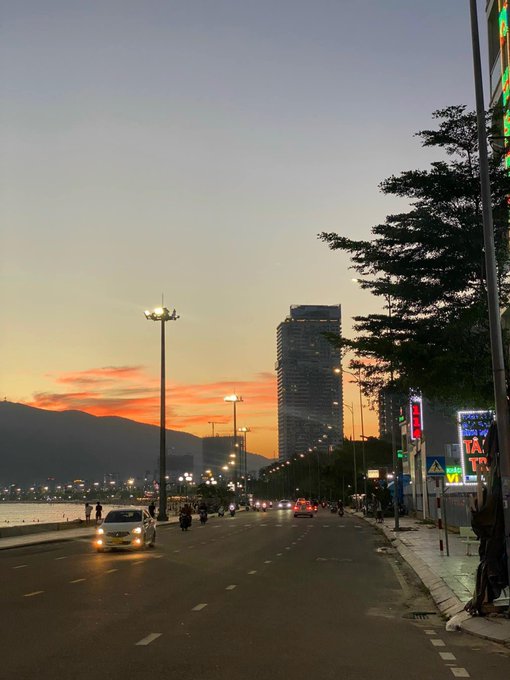 A coastal road at dusk with streetlights, cars, and a tall building in the distance, with the sun setting behind mountains across the water.