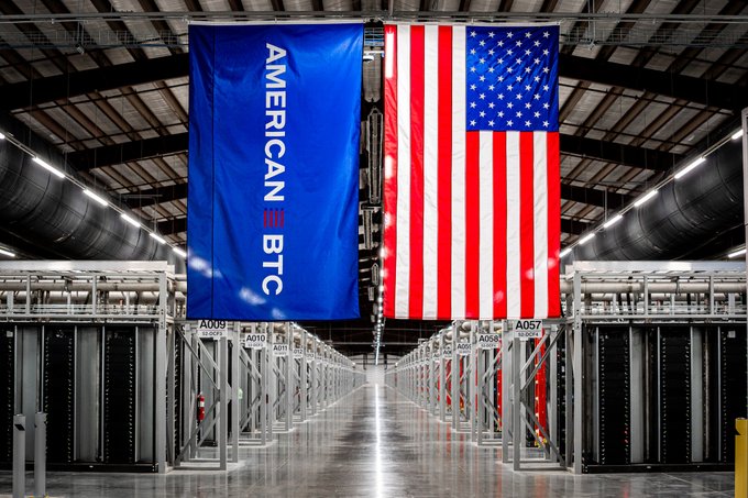 Two large flags—a blue 'AMERICAN BTC' banner and an American flag—hang from the ceiling of a large server/data center facility with rows of computer racks on either side.