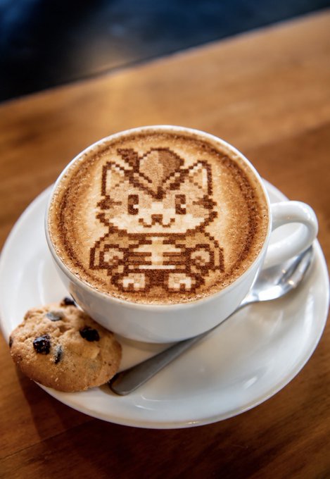 A cup of cappuccino features latte art depicting a cute cartoon cat character, served alongside a chocolate chip cookie on a white saucer.