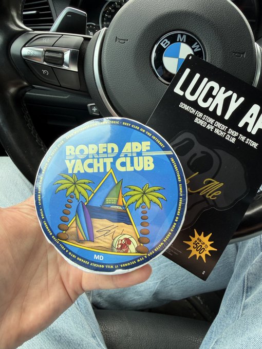 A hand holds a circular 'Bored Ape Yacht Club' sticker and a 'Lucky Ape' scratch card inside a BMW vehicle.