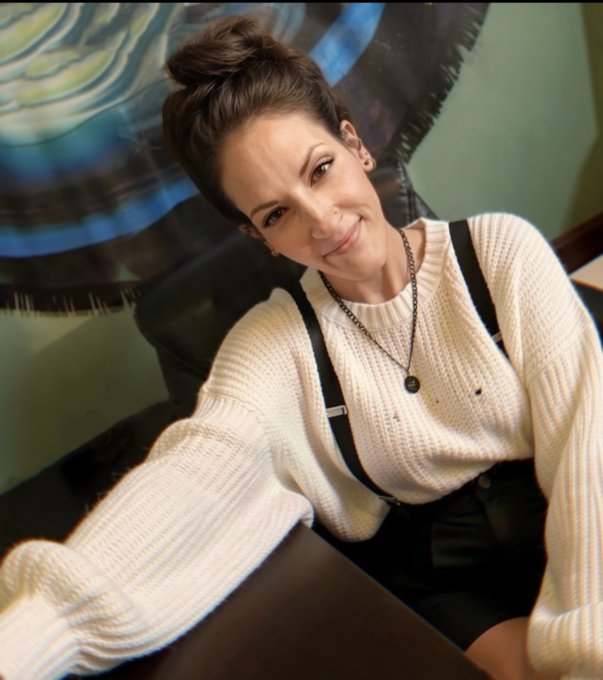 A woman with her hair in a top knot smiles for a selfie wearing a cream sweater and suspenders, projecting a confident, playful vibe that mirrors the witty, self-deprecating humor of the post joking about not being able to afford a drug habit.