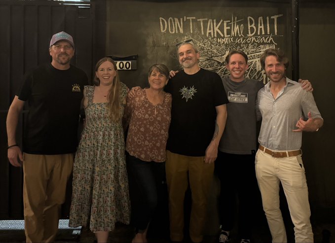 A group photo of six people smiling and standing together in front of a wall with a mural that reads 'DON'T TAKE the BAIT - Marina del Rey' featuring shark illustrations.