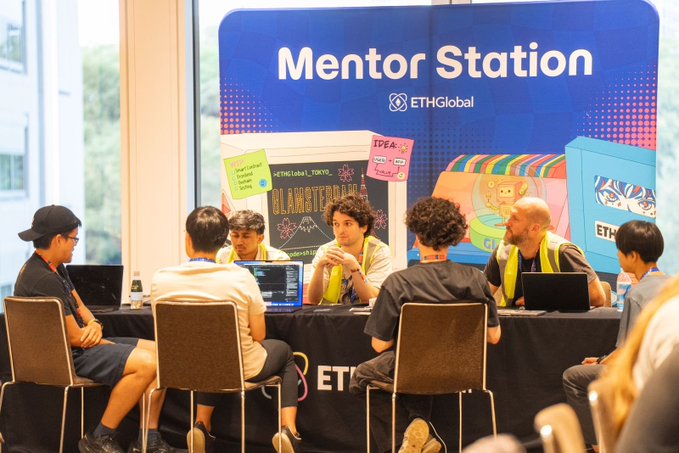 A group of people sit around a table at an 'ETHGlobal Mentor Station' booth, with signs including 'BLAMETEDA' and 'IDEA' visible, discussing and using laptops.