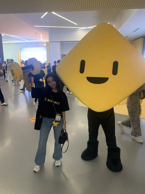 A woman wearing a black "BINANCE" t-shirt and lanyard poses next to a large yellow mascot holding a "1000" sign at the Binance Blockchain Week event. The image captures her role as a Binance Angel and community representative celebrating Binance's 9th anniversary.