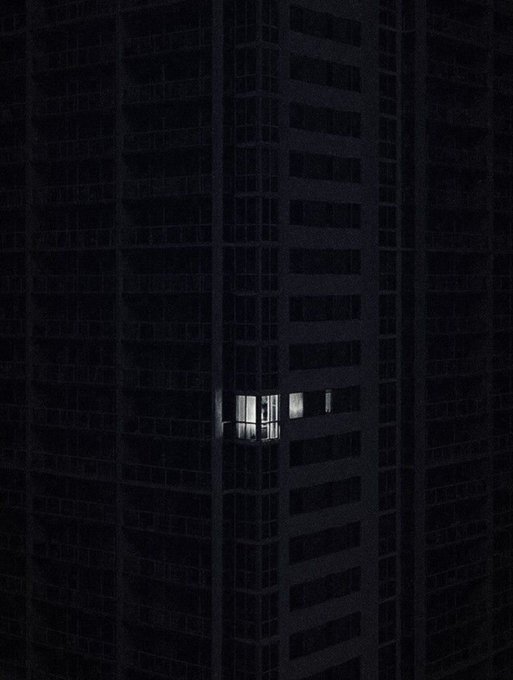 A single lit window glows in an otherwise pitch-black high-rise, suggesting one lone tenant still awake among countless darkened units. The image visually reinforces the caption "They sleep, while you grind for the bag," symbolizing late-night hustle amid a sleeping world.