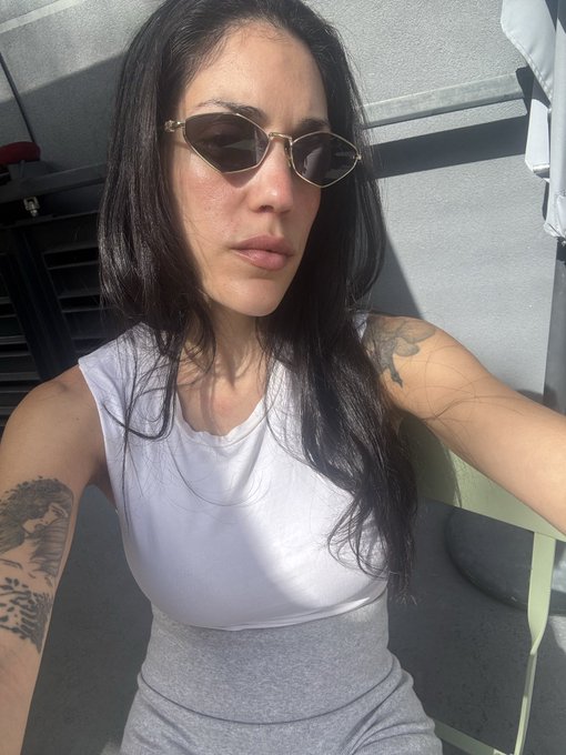A selfie of a woman with long dark hair wearing small oval sunglasses, a white t-shirt, and gray bottoms, with visible tattoos on her arm, seated outdoors in sunlight.
