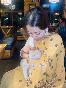 A young woman in a yellow floral traditional outfit smiles down while holding a small white cat in a dimly lit restaurant or lounge setting.