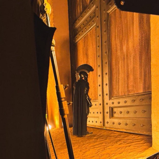 A figure dressed as a Roman centurion, wearing a plumed helmet and dark cloak, stands before a large, ornately studded wooden door in a warmly lit, sandy-colored setting.