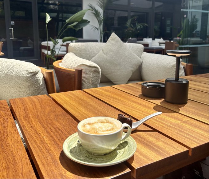 A frothy cappuccino in a white cup on a green saucer, sitting on a wooden table in a sunlit cafe setting with beige cushioned chairs and a small lantern.