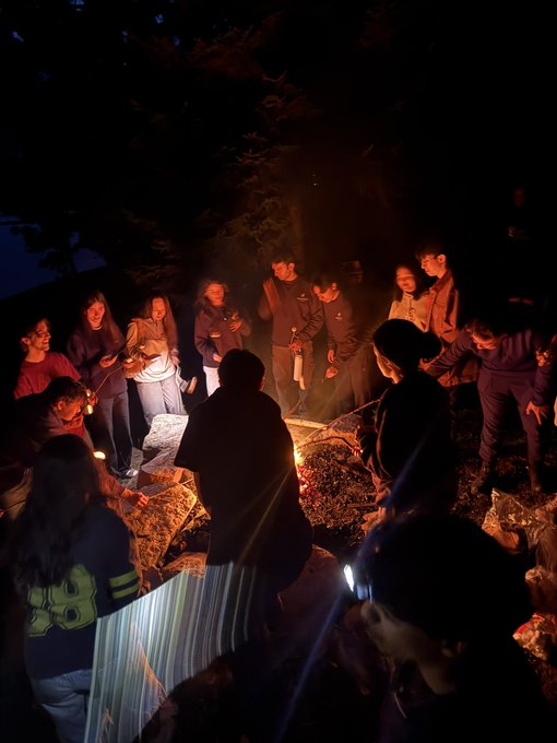 A nighttime outdoor gathering where a group of people stands around a campfire, illuminated by the warm glow of flames and a flashlight.