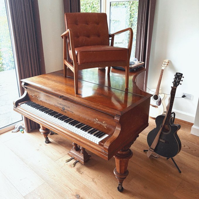 A wooden upright piano with a brown leather chair sitting on top of it, alongside two electric guitars on stands in a room with hardwood floors and large windows.