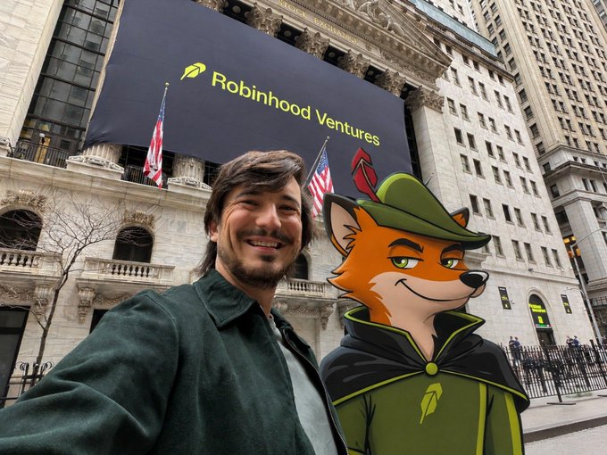 A man takes a selfie with a fox mascot wearing a green hat in front of the New York Stock Exchange, where a large digital banner displays "Robinhood Ventures" with the company's logo, tying into the post's message about Robinhood's mission to democratize finance for all and its connection to $FOX.
